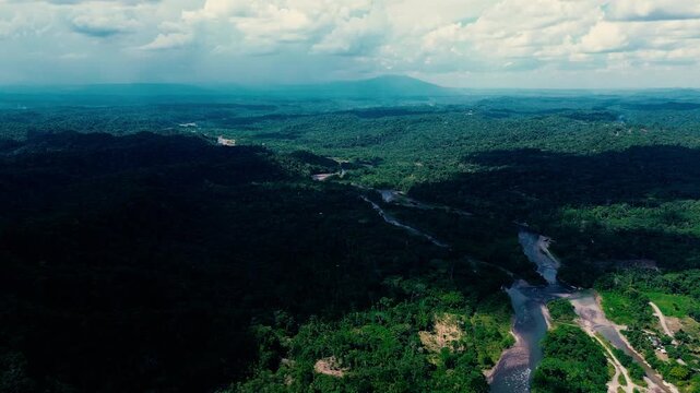 Aerial drone footage shows a small settlement in Ecuador's Oriente, where roads and scattered buildings open into dense tropical forest. Forested hills and a broad, cloud-filled sky frame the town and