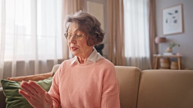 Cheerful senior Caucasian woman in glasses talking during video call while sitting on sofa. Smiling elderly female communicating online with family or friends from comfortable home. Saying bye.