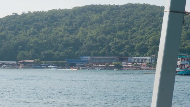 Point of view from a moving passenger ferry boat cruising past Thonglang Beach towards Tawaen Pier on Koh Larn in Pattaya Thailand showing calm ocean water anchored tour boats and lush green mountains