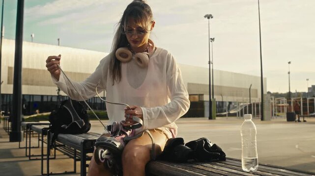 Young woman unlaces inline skates while sitting on wooden bench. She wears sunglasses and headphones preparing for active outdoor rollerblading session during warm summer day.