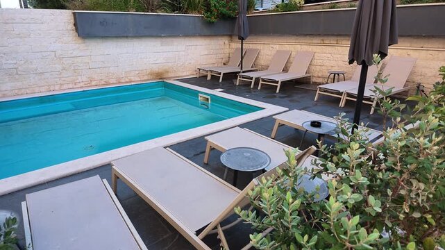 Outdoor swimming pool surrounded by beige sun loungers, umbrellas, Mediterranean plants and stone paving