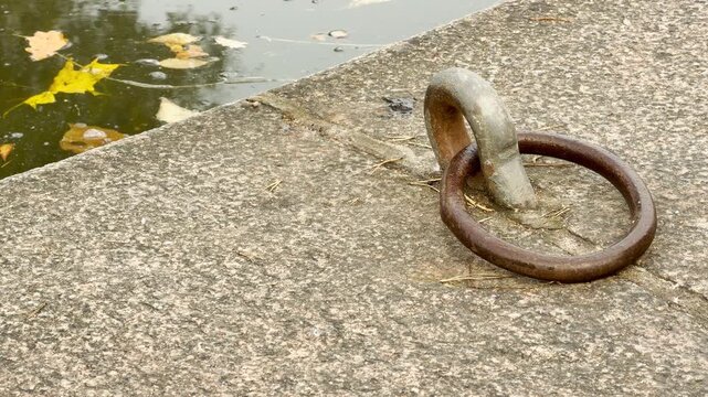 A rusty metal mooring ring rests on a granite pier beside still lake water. Fallen yellow autumn leaves float gently, creating a quiet and nostalgic mood. This peaceful spot is Catherine Park, Pushkin