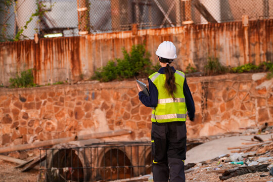 Environmental compliance engineer walking toward an outfall below a security fence and storage tank, woman seen from behind in hard hat and hi-vis vest in Thailand