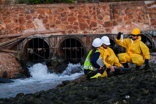 Technician in yellow hard hat raising a sample vial to the light while a woman engineer and bearded colleague crouch beside him at a wastewater outfall in Thailand