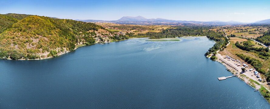 Aerial panorama ideal for tourism and travel - wide lake surrounded by forested hills, small dock, and mountain backdrop in summer - Balkans.