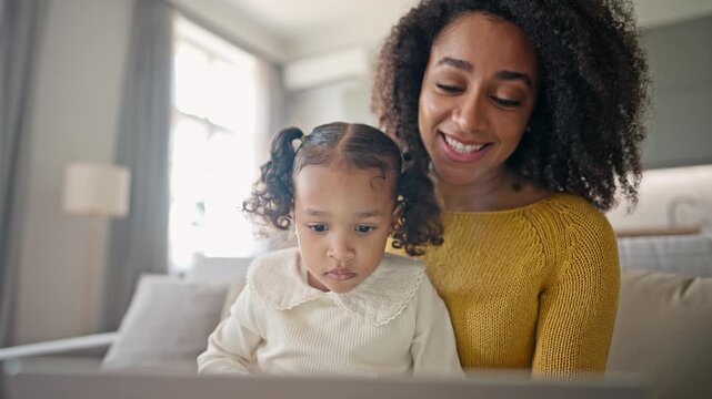 Happy African American mother sitting closely with little cute daughter and supporting her during learning activity. Caring parent smiling while focused child developing skills at home.