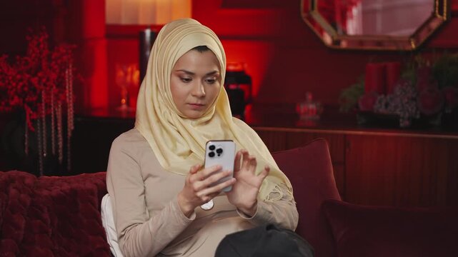 Puzzled Middle Eastern woman in hijab shopping online using smartphone, choosing