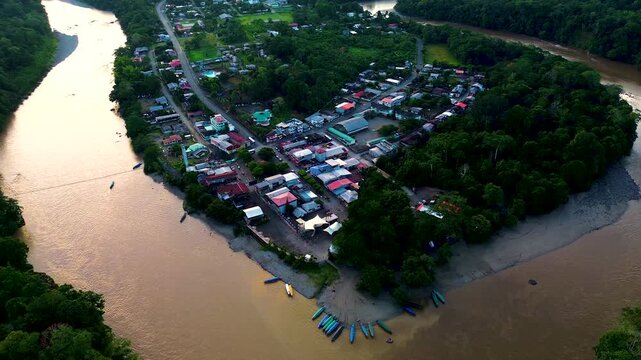 A broad aerial panorama shows a muddy river curving through dense tropical forest and past the riverside town of Misahualli, Ecuador. Green hills and distant ridgelines sit beneath low clouds, with ro
