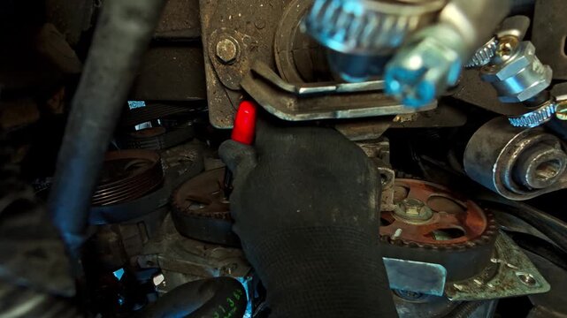 A car mechanic in a glove uses a red pen to draw a timing mark on the camshaft sprocket and belt during engine repair, close-up.