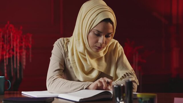 Arab female student in hijab preparing for exam, reading and taking notes