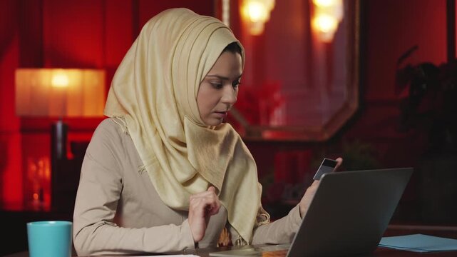 Young Arab woman in hijab entering card details on laptop, shopping online