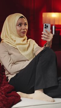Relaxed Muslim woman in hijab talking with friend on phone video call at home