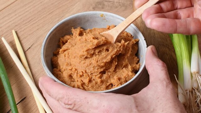 A hand scoops a spoonful of shiro miso paste from a bowl beside fresh scallions and chopsticks.