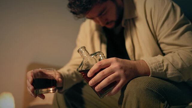 Solitary adult man grips whiskey glass and liquor bottle on dark bed. Profound sorrow and grief