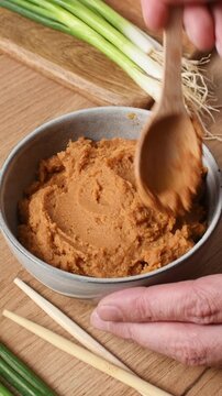 A wooden spoon spreads and smooths shiro miso paste in a bowl near scallions and wooden chopsticks.