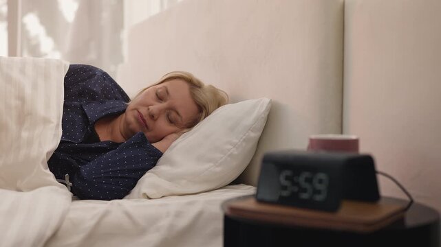 Exhausted middle-aged woman turns off alarm clock and goes back to sleep