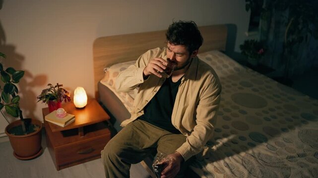 Alcohol dependency drives sorrowful adult male to sit on bed with glass. Acute mental depression