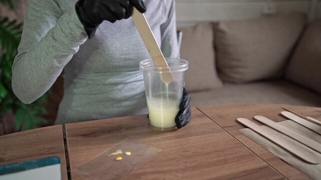 Closeup hands in black gloves stirring pale resin in clear plastic cup with wooden stick on wooden table, yellow pigment nearby in indoor craft workspace