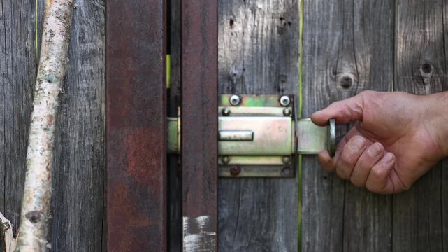 Caucasian man unlocks rural wooden fence gate with sliding metal bolt latch, opening backyard entrance smoothly into sunny green garden area during tranquil day outdoors with natural light