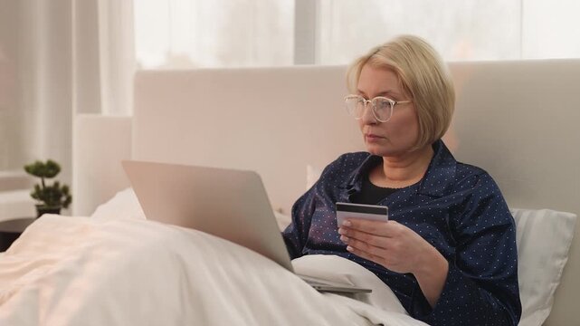 Senior woman in glasses enters credit card details while shopping online in bed