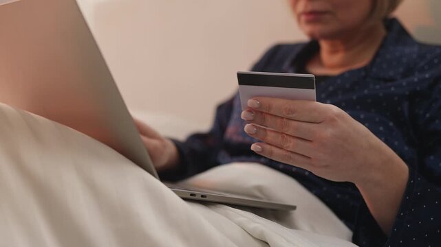 Mature woman shops online in bed, entering credit card details on laptop