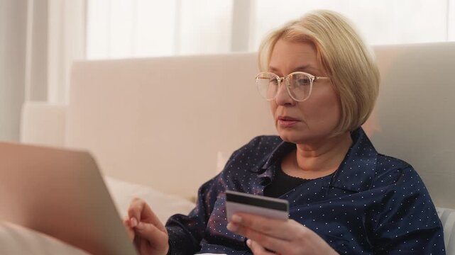 Senior woman enters credit card details on laptop while shopping online in bed