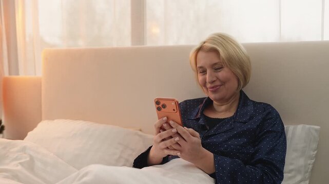 Happy middle-aged woman watches funny videos on phone while relaxing in bed