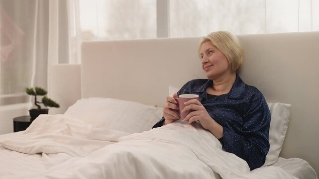Happy middle-aged woman enjoys morning coffee while relaxing in comfortable bed