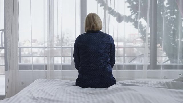 Depressed middle-aged woman sits on bed looking out window, feeling lonely