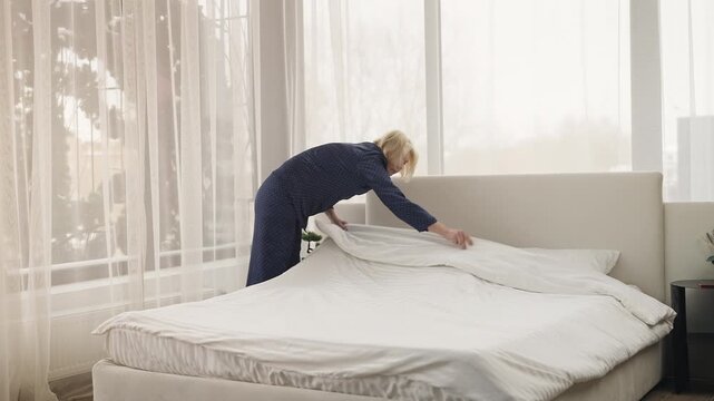 Tidy middle-aged woman makes her bed in morning as part of daily home routine