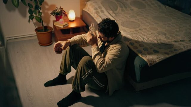 Sitting on floor beside bed troubled adult male clutches liquor bottle. Deep despair and sorrow