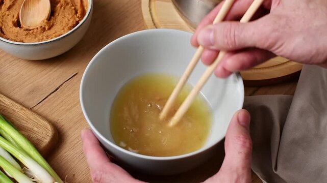 Chopsticks stir shiro miso paste into broth in a bowl, gradually dissolving it into warm broth.
