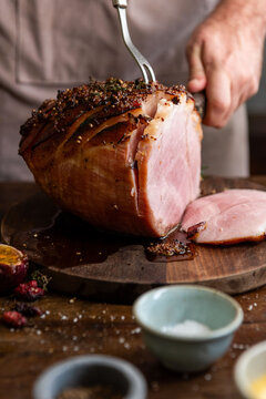 Person carving a glazed ham on a wooden board with spices nearby.