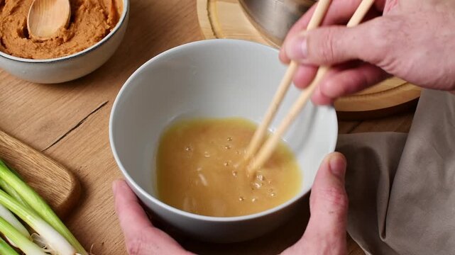 Chopsticks stir shiro miso into broth in a bowl, blending the paste into a smooth, cloudy mixture.