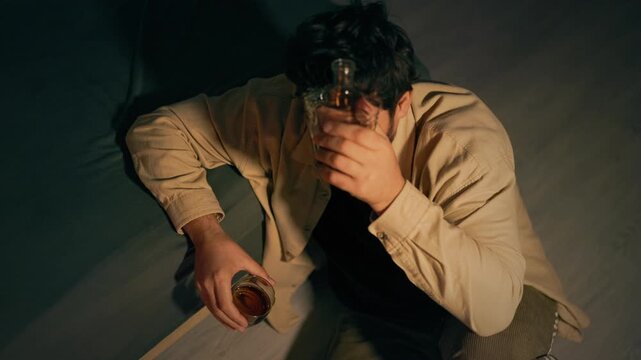 Sorrowful adult man swallows intoxicating drink on floor near bedside. Severe emotional breakdown