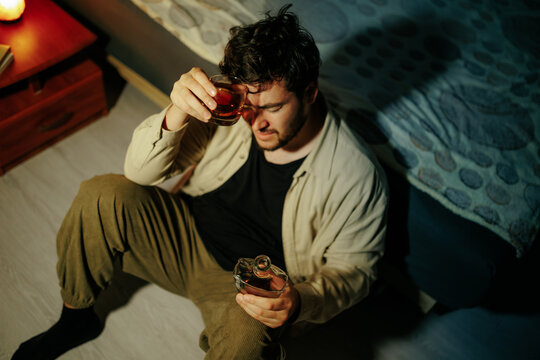 Alcohol dependency drives anxious man to sit on home floor with bottle. Life crisis despair