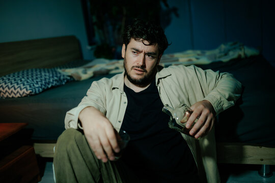 Stressed man battles acute anxiety holding alcohol bottle. Dark bedroom life hardship pain
