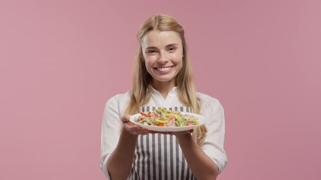 Beautiful young female cook in apron holds fresh appetizing salad,smells its aroma and invites to taste it. Healthy food concept for recipes