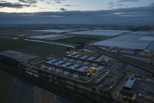 Aerial view of a modern hyperscale data center facility illuminated at dusk, surrounded by vast agricultural fields and greenhouses in the Netherlands.