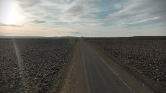 Aerial drone shot moving forward following a white camper van driving along a remote straight asphalt road through a vast barren rocky desert landscape under a cloudy sunset sky.
