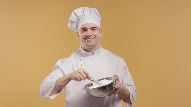 Professional male chef in white chef jacket and hat whisks fluffy egg white cream. Culinary preparation concept for baking, desserts