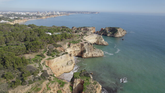 Aerial view of Ponta Joao de Arens featuring rugged limestone cliffs, turquoise Atlantic waters, coastal hiking trails, and pine trees along the coastline in Alvor, Faro, Portugal.