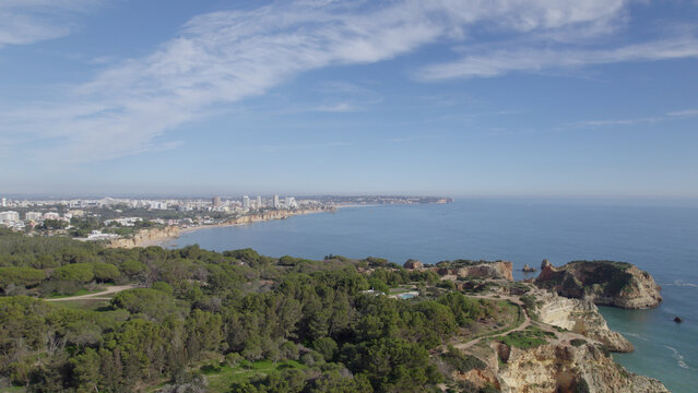 Aerial view of Ponta Joao de Arens with its rugged cliffs, lush pine forests, and the coastline stretching towards the town of Alvor, Faro, Portugal.
