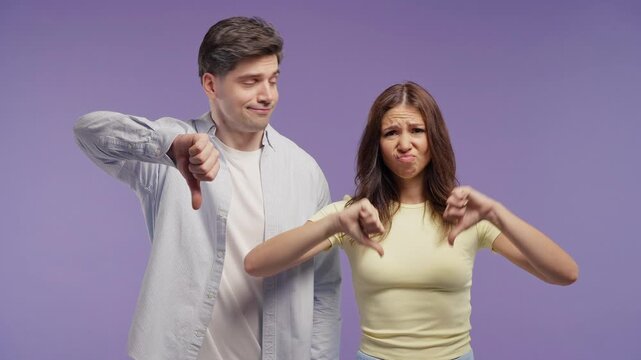 Unhappy couple of woman and man condemns with dislike sign. Friends expressing discontent with showing thumbs-down gesture on violet studio.