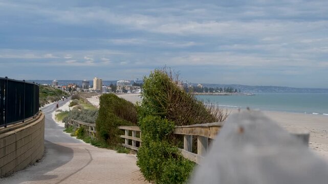A scenic view of West Beach coastal path looking toward the Glenelg skyline,featuring a shared walking trail, protective dune fencing, native vegetation, sandy shoreline. Adelaide, South Australia