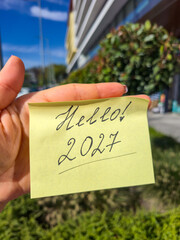 Hand holding a yellow sticky note with text Hello 2027 against a green bush and a building. Concept of welcoming the New Year 2027, greeting and new start, grainy film texture, high quality photo © Татьяна Швец