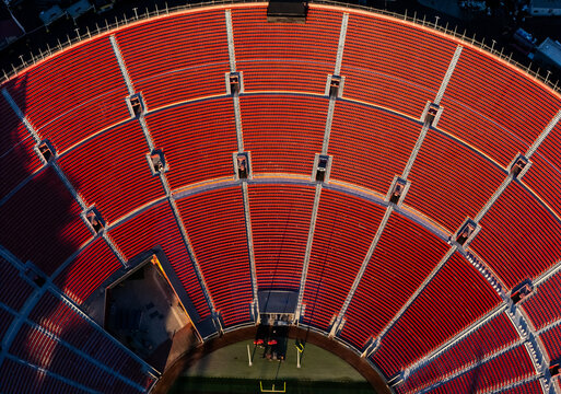 Aerial view of LA Memorial Coliseum showing the vast rows of empty red stadium seats under the warm light of the golden hour in Los Angeles, California, United States.