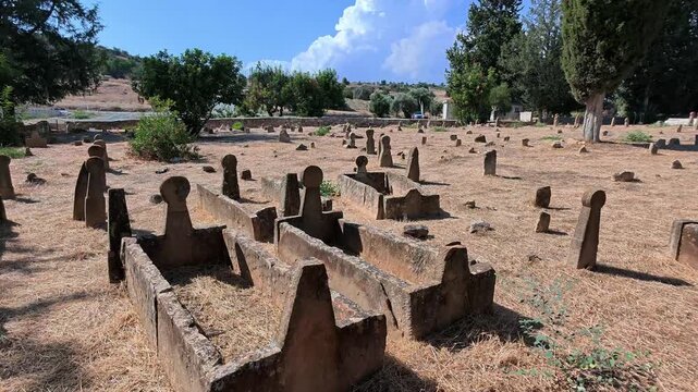 Tochni Turkish Cypriot cemetery in Cyprus with old stone graves and tombstones scattered across dry ground, surrounded by cypress trees and hills beneath a blue summer sky