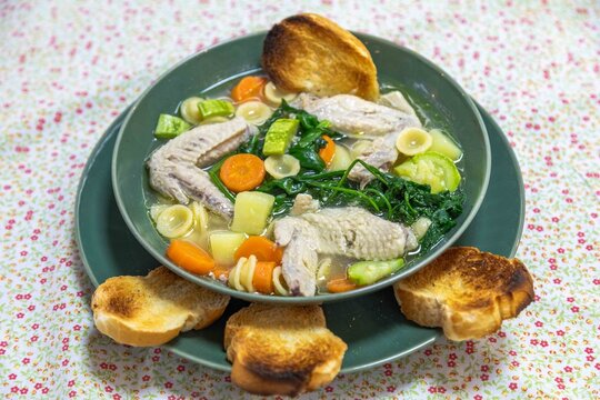 Homemade chicken wing soup with carrots, chayote, potato, spinach and small pasta shells in a clear broth, served in a green ceramic bowl with toasted bread on a floral tablecloth.