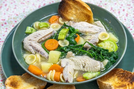 Homemade chicken wing soup with carrots, chayote, potato, spinach and small pasta shells in a clear broth, served in a green ceramic bowl with toasted bread on a floral tablecloth.